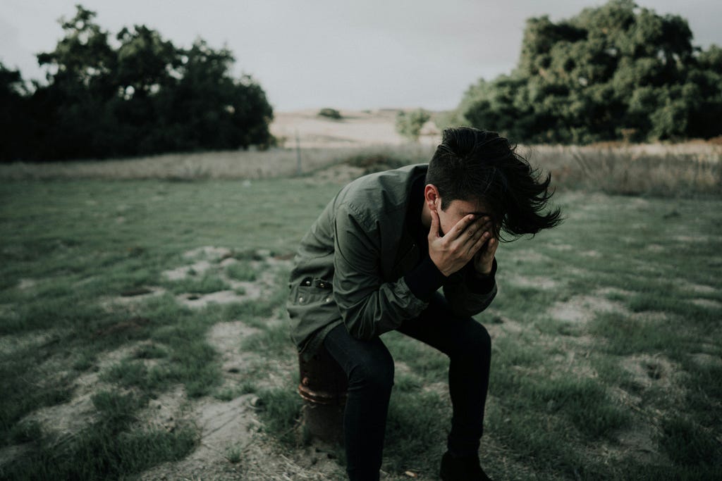 Photo of a young man crying, dressed in a green parka and black pants, he is sitting on a log. Located on a natural patch of grass, in the background you can see a meadow with trees and dry plants similar to cut wheat.