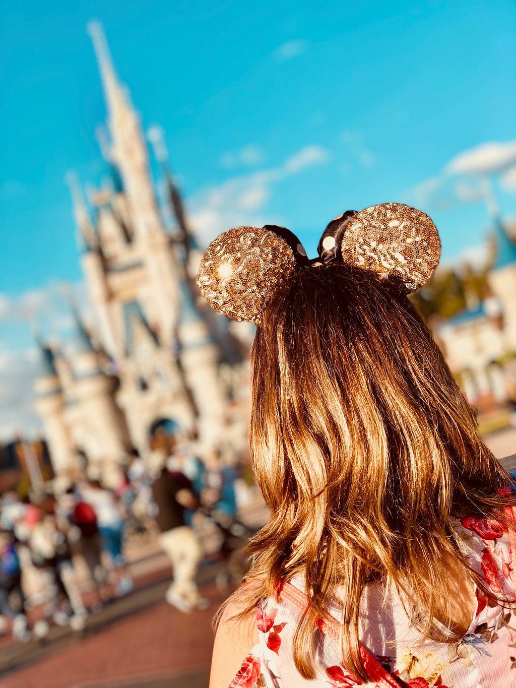 We see the back of a woman’s head in gold, sparkly Minnie Mouse ears as she walks toward Cinderella Castle at the centre of a Disney theme park — alt text to boost Medium stories.