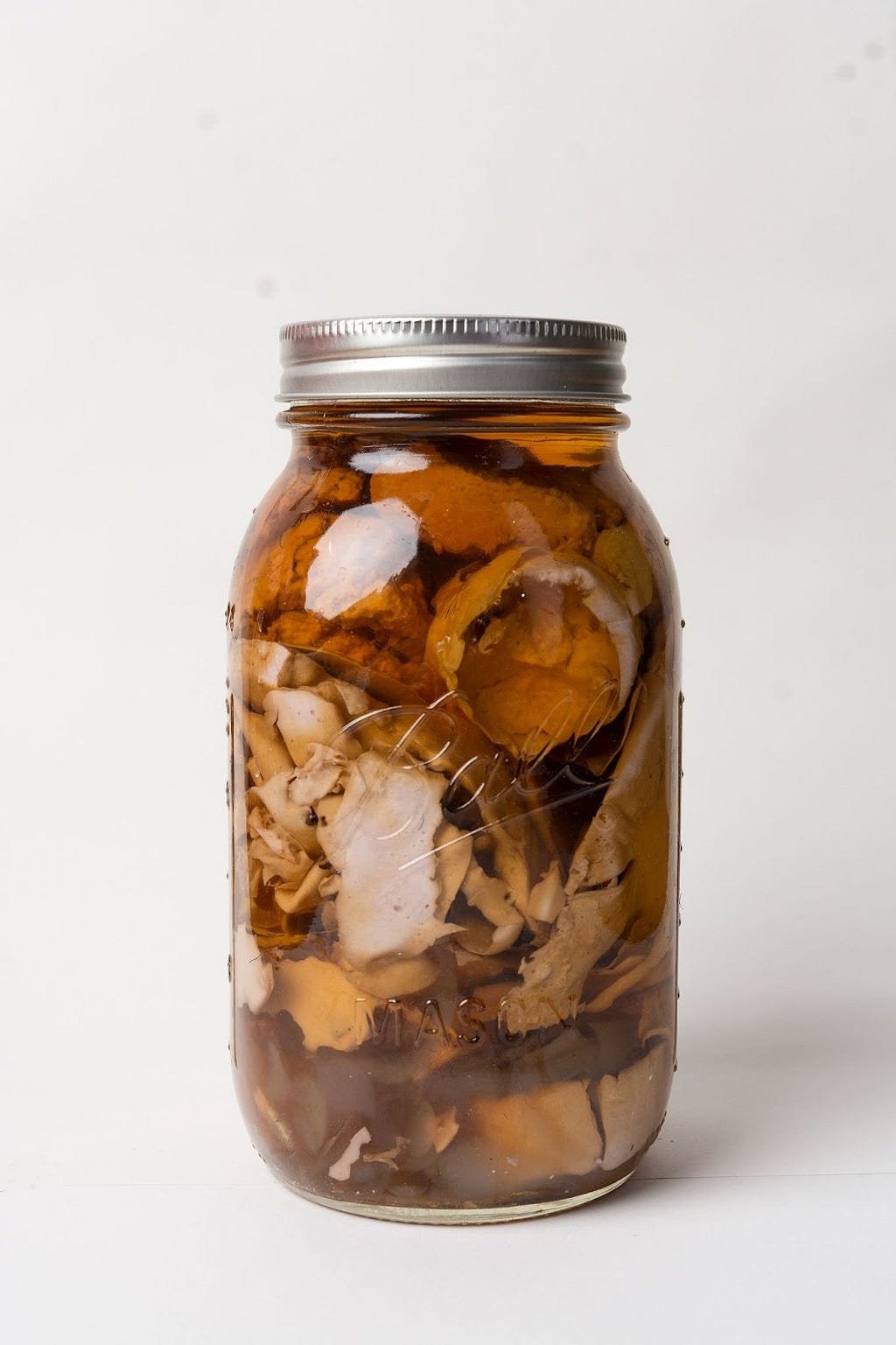 A glass jar with a metal lid is inscribed with the text ‘Ball’ and ‘mason’ and sits on a white background. The mason jar is full of pale purple alginate remnants and bioplastics surrounded by a light brown liquid. The jar represents preserved memories.