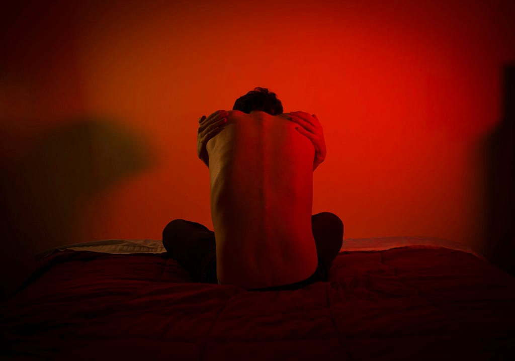 A photo of a shirtless man sitting with his back facing the camera. He’s hunched over and has his arms wrapped around himself. The lighting is red, giving an anxious feel to the image.