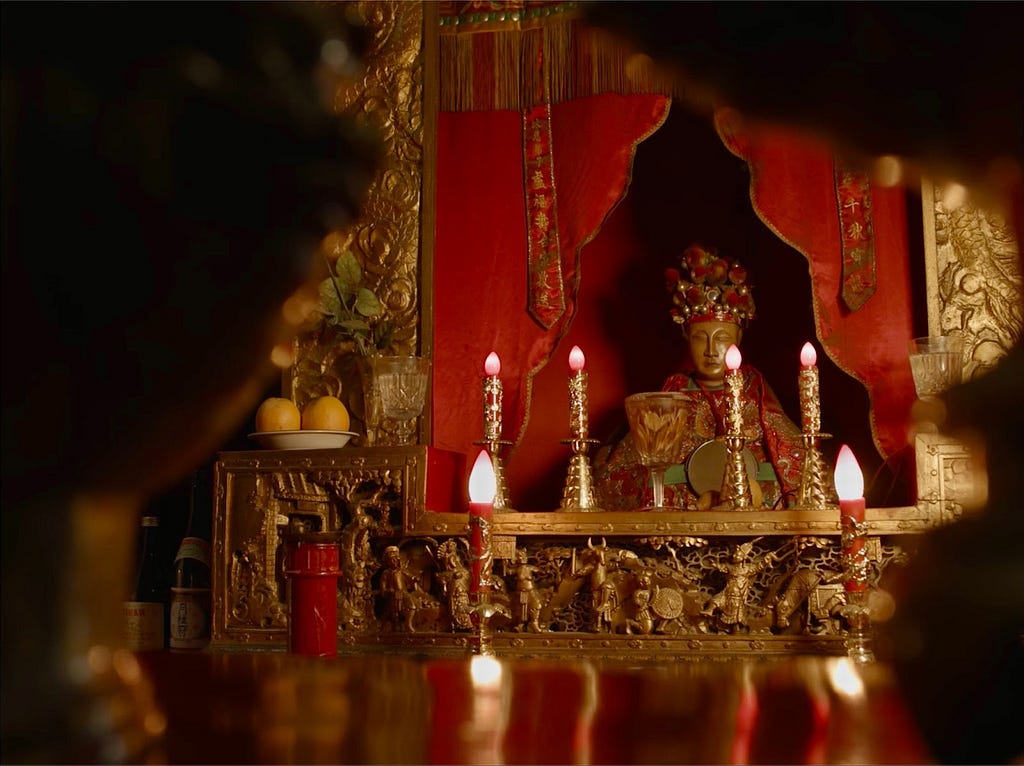 Gold flameless candles adorn the red altar to Tam Kung. The shot is partially silhouetted in the shadows of a curving shape.