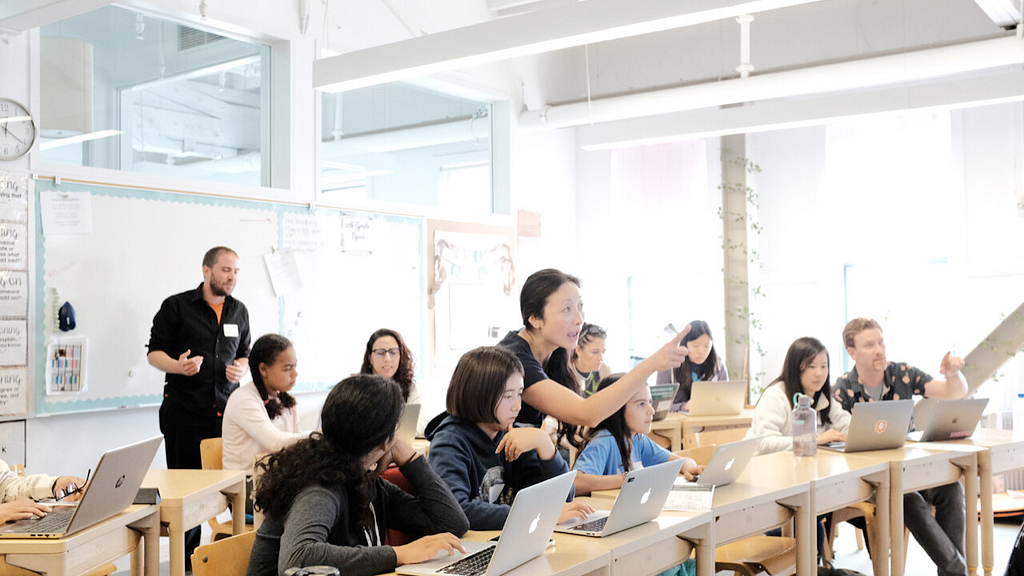 A photograph of a classroom with twelve people. The students sit at desks with laptops and several people gesture and point while speaking to them. Angi is at the center, pointing to the right to show a student something.