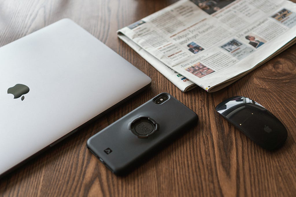 A laptop, phone, wireless mouse and newspaper on a wooden desk.