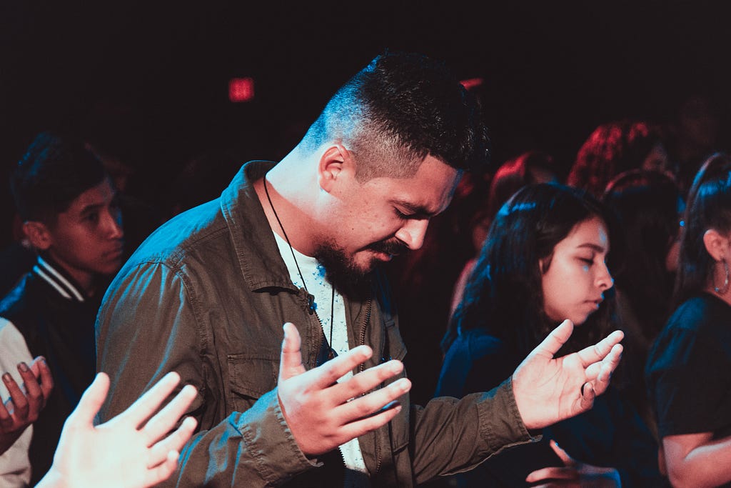 Group of Middle Eastern descendent persons standing and praying with man closest having head bowed and hands lifted up to chest-level; a woman with her eyes closed in prayer stands next to him.