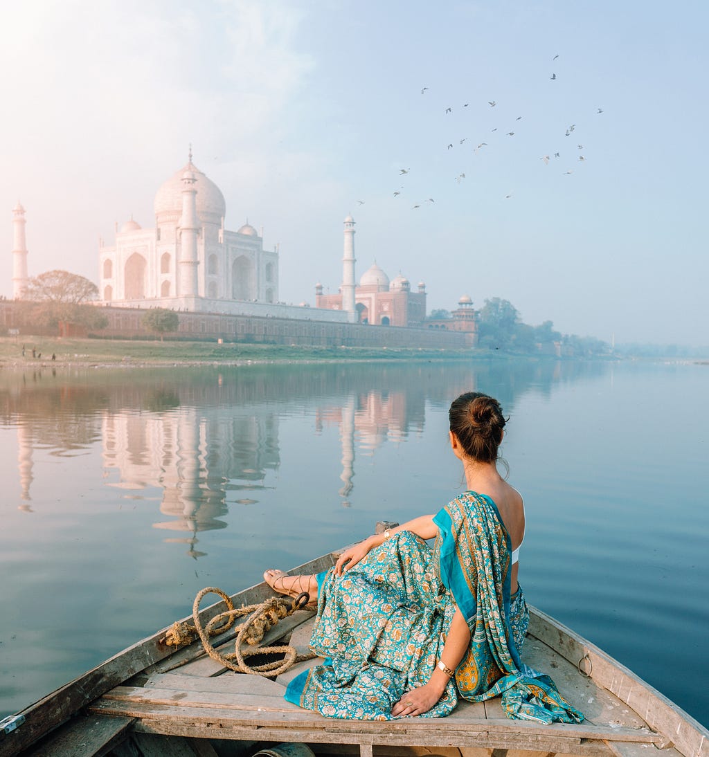 A woman sits on the front of a boat in an Indian sari and looks out at the majestic Taj Mahal that appears in the distance.