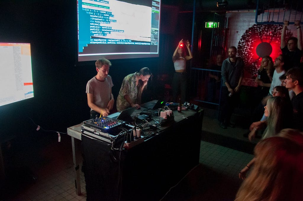 A photograph of a live performance, with two people standing at a table on stage with laptops and a sound mixer. Behind them is a projection of the screen. An audience faces them.
