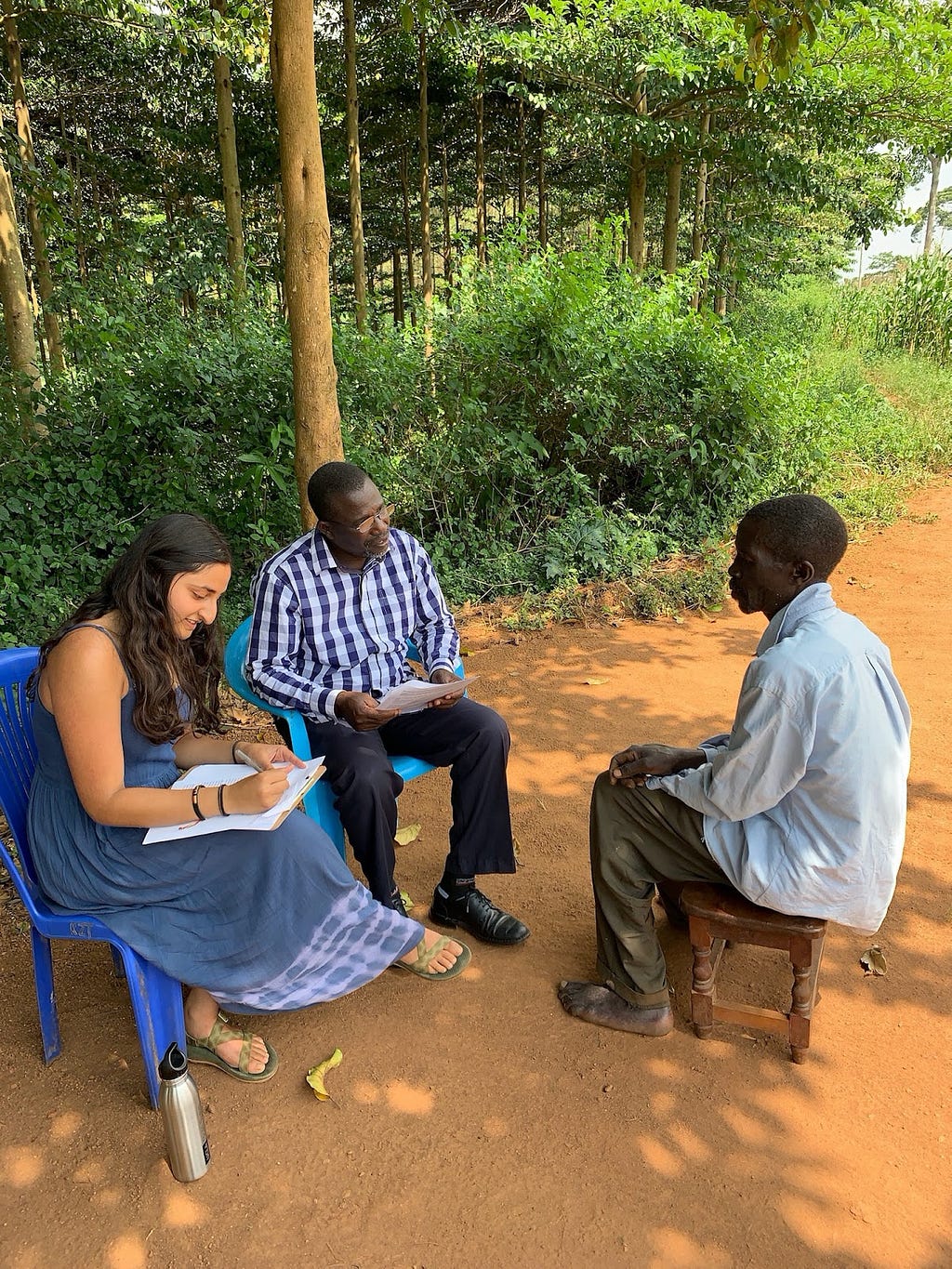 Three people sit on chairs on a sandy path in a rural area, two talking and one taking notes.