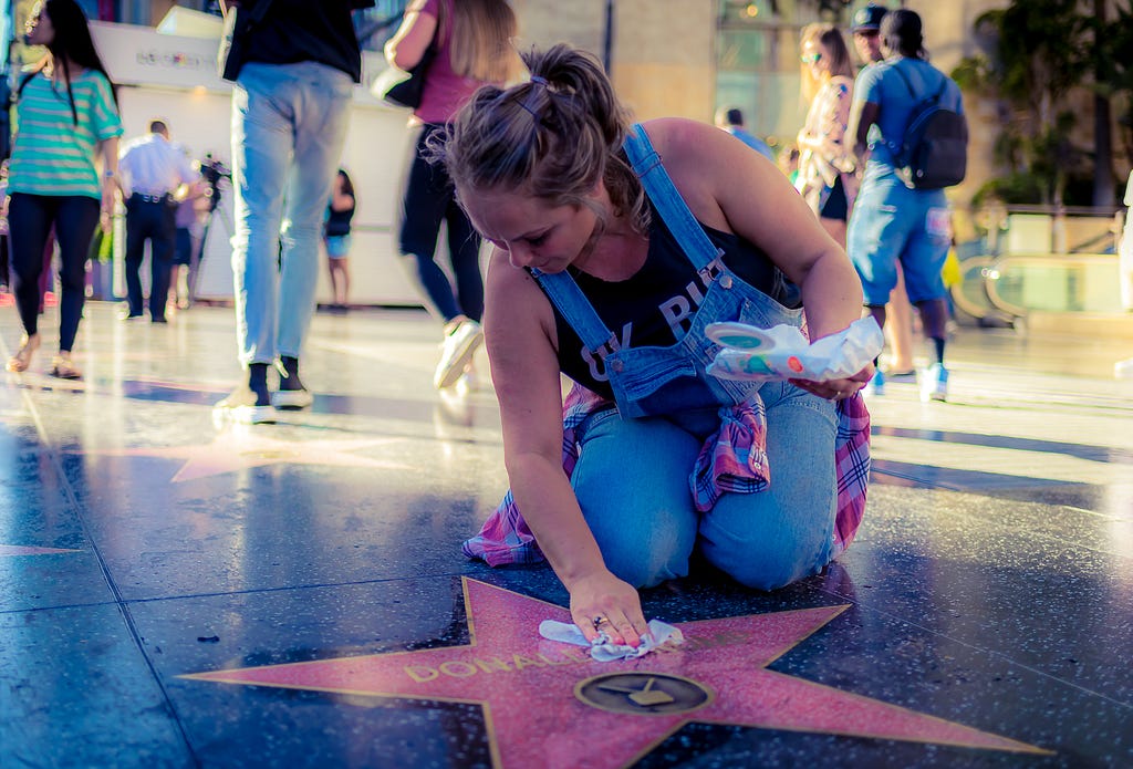 a woman in denim overalls kneeling at Donald Trump's star on Hollywood Blvd and cleaning it with baby wipes