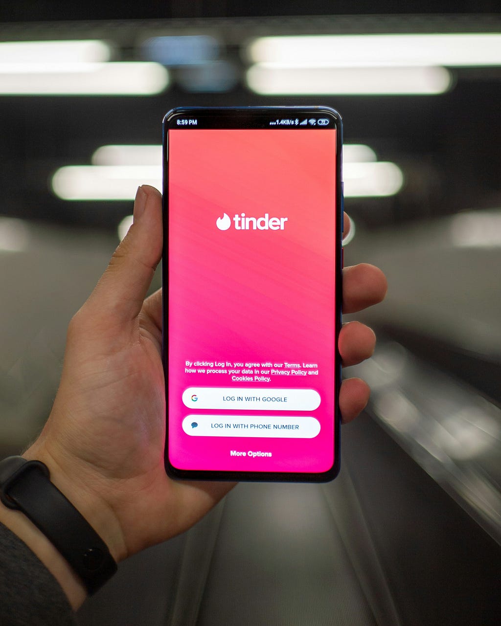 Uma mão segurando um smartphone com a tela de login do Tinder aberta, com opções de logar via Google ou número de telefone. O fundo, atrás do smartphone, está borrado.