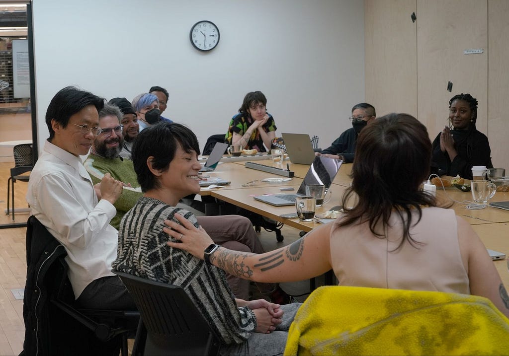 A behind-the-scenes image of our board of directors and advisors, staff, and community members at NYU ITP. Around ten people gather by a table while working on their laptops. Lauren Lee McCarthy is at the front of the image, laughing while someone pats their shoulder.