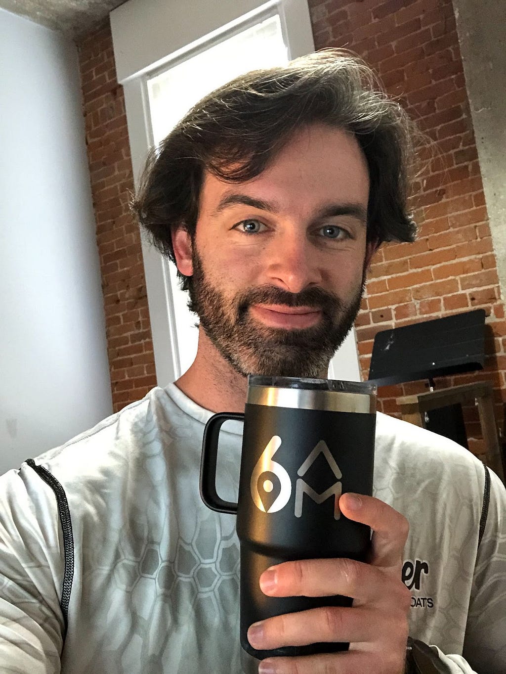 A bearded man smiles as he holds up a black travel mug with 6AM logo branding.