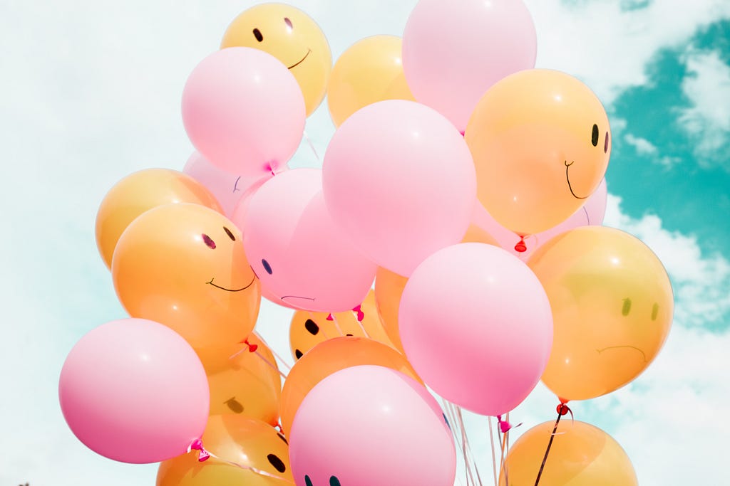 A close up of a cluster of balloons, pink and yellow, attached all with various smiley faces painting onto them atop a very blue sky with white clouds.