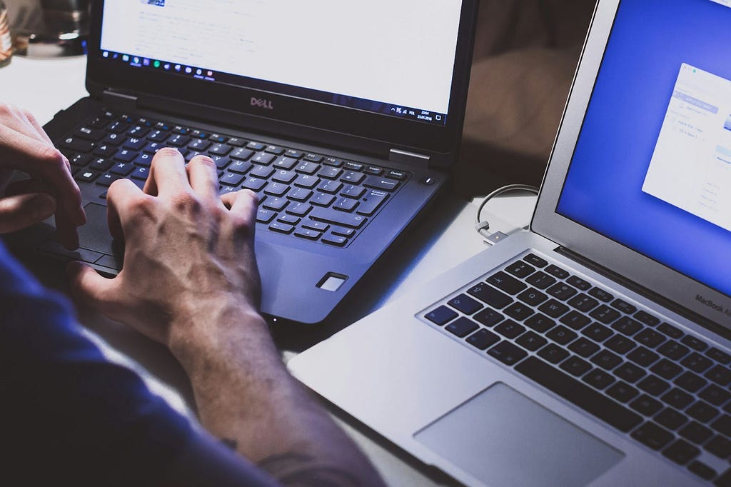 Somone sitting in front of two laptops, typing on the one on their left.