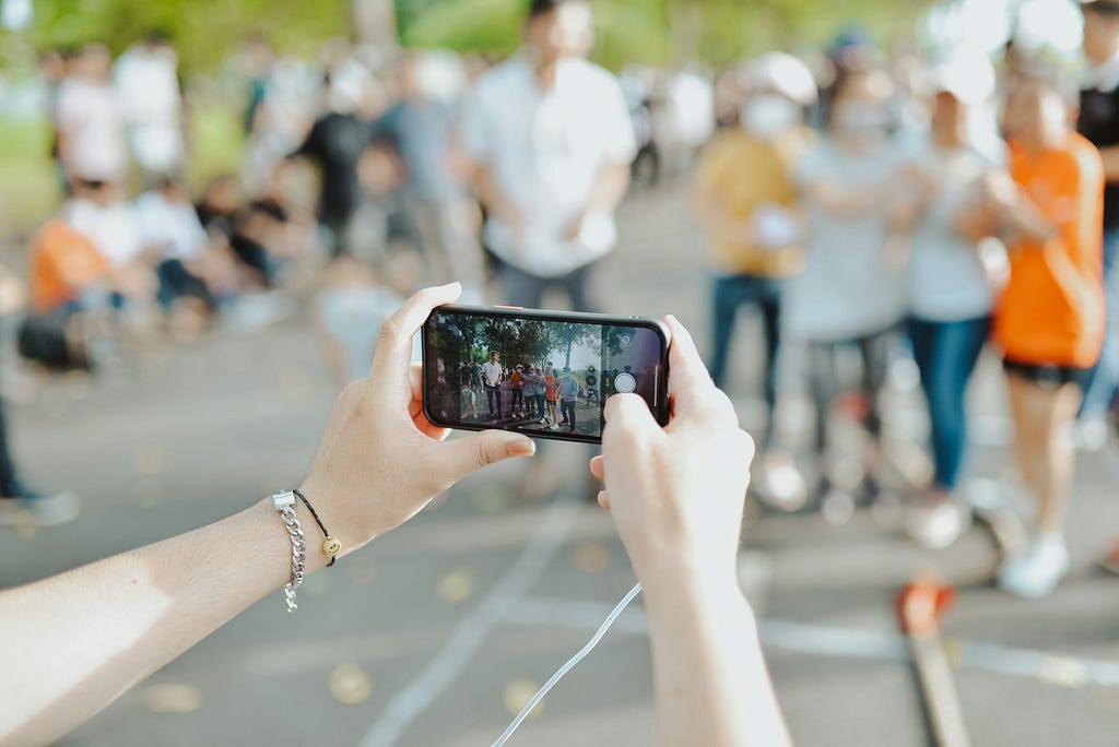 The image shows someone holding a smartphone and taking a photo of a group of people outdoors.