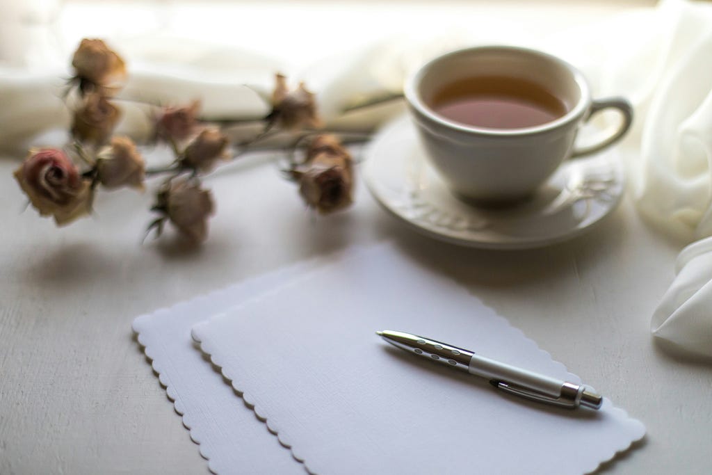 Two pieces of blank stationery and a pen rest on a white table, with a cup of tea in a teacup rests on a saucer beside a stalk of dried pink flowers