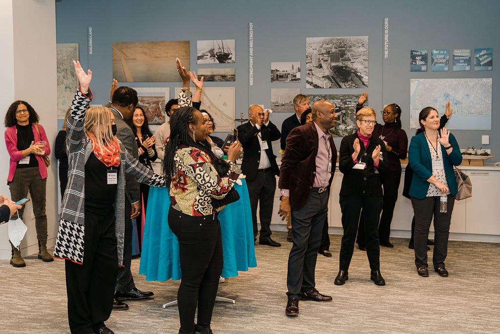 Candid photo of 1–2 dozen people standing in a museum wearing business clothes. They are smiling and raising the hands.