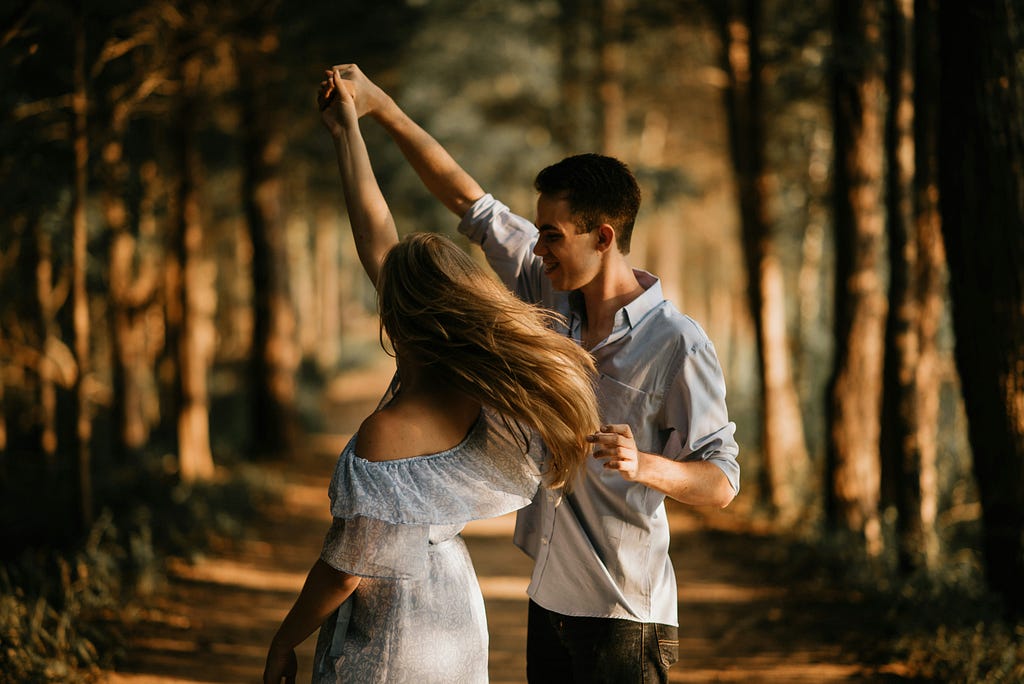 A color photo of a young girl and boy dancing along a wooded lane outside. The sun filters through casting shadows on the dirt road. The young man has the woman by her hand and is twirling her. Her long hair if flying, her white dress if off her shoulders. He holds her hand high as he spins her. Their bodies are not touching — she’s spinning, her face can’t be seen. He looks almost as if a puppeteer pulling her strings.