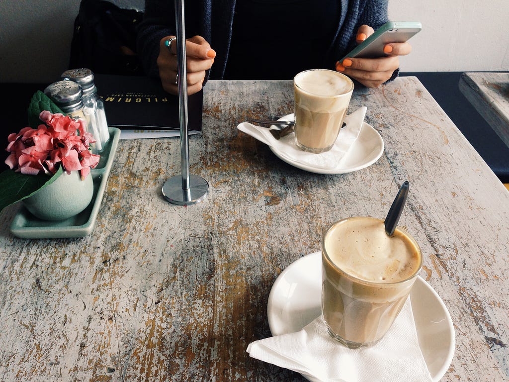Image of a square, 2-top café table, with two cappuccino’s and a person seated on one side holding a cell phone.