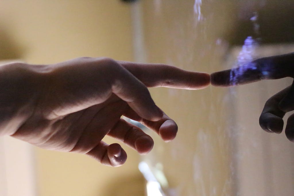 Photograph of a left hand with index finger extended to the right and touching its reflection in a glossy surface.