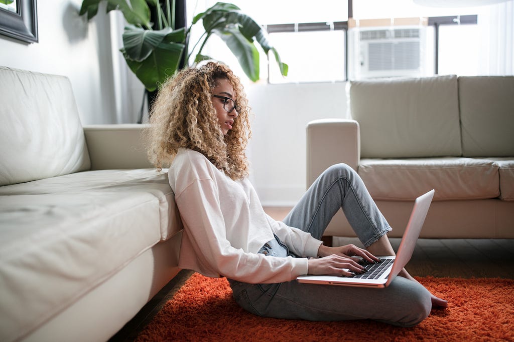 A woman with curly hair sitting on the floor of an apartment typing on her laptop.