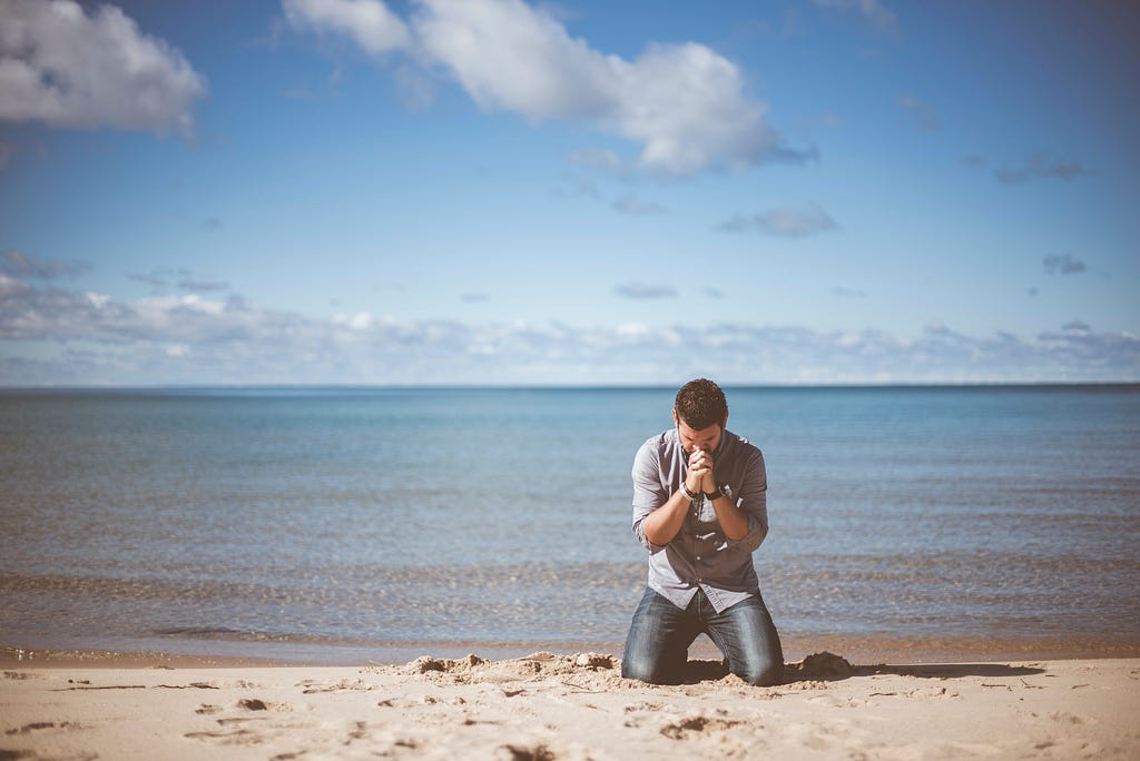 A man kneeling in prayer in front of a body of water.