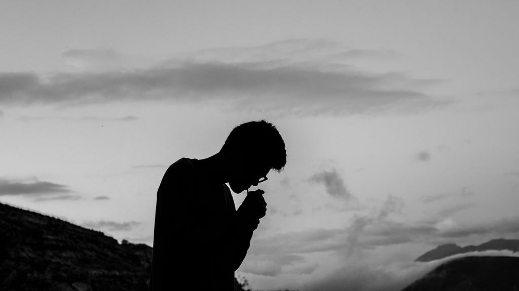 A silhouette of a person lighting a cigarette and looking down a mountain.
