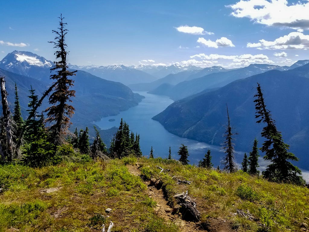 Atop a mountain overlooking the water, this scene from the point of view of a hiker who is looking out into the horizon upon reaching the heights he/she was hiking for. There are trees in the foregrounds and clourds trailing the right corner. Shades of blue, green, and browns are shown.