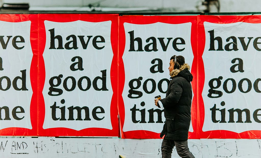A man in a black winter coat walks past posters with bold black text on a white background reading “have a good time” repeated across a red border.