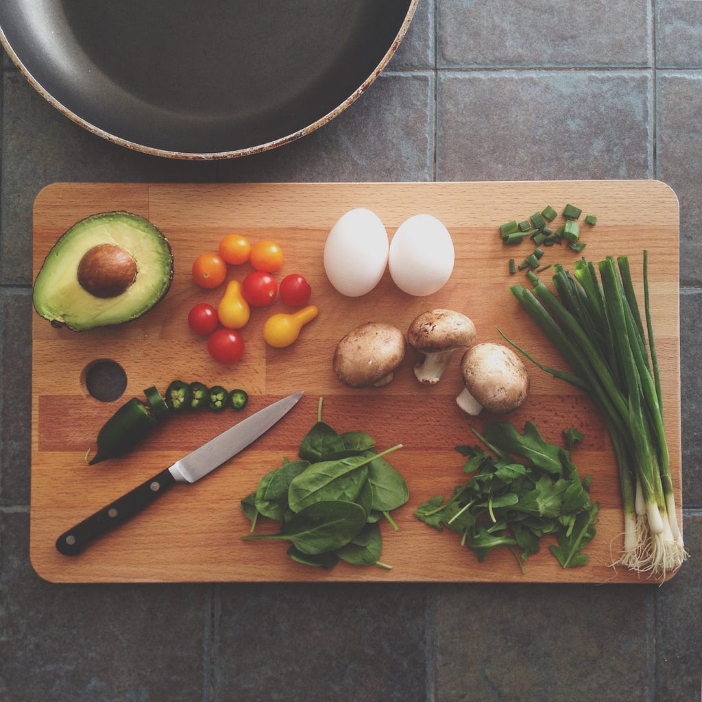 Foto de uma tábua de corte de alimentos, com verduras e legumes espalhados por cima.