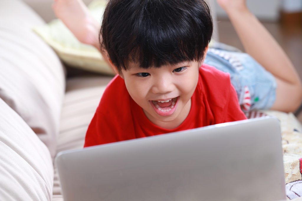 A photo of a young boy lying on a couch, talking into a laptop computer.