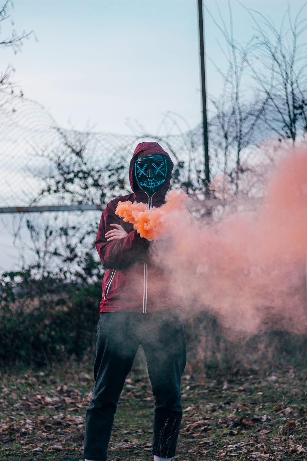 Person in a hood and face mask with eyes crossed out, in a cloud of orange smoke