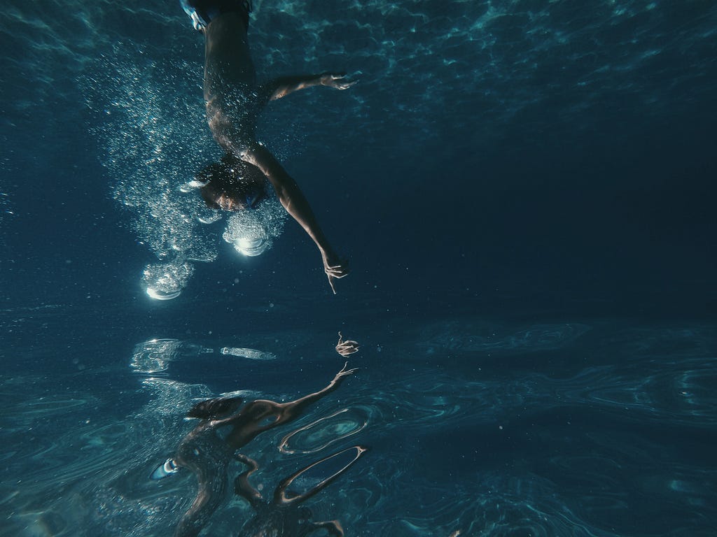 A man wearing swimming goggles reaches out to the water's surface from underwater.