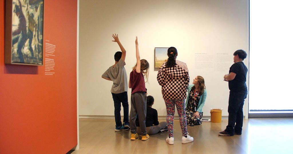 An educator kneels on the floor in a gallery with young students standing and sitting around them during an Instill lesson. Some students raise their hands