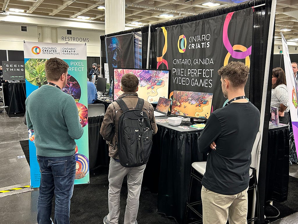 A group of convention-goers stand inside a booth. One of them is playing a video game as the others observe.