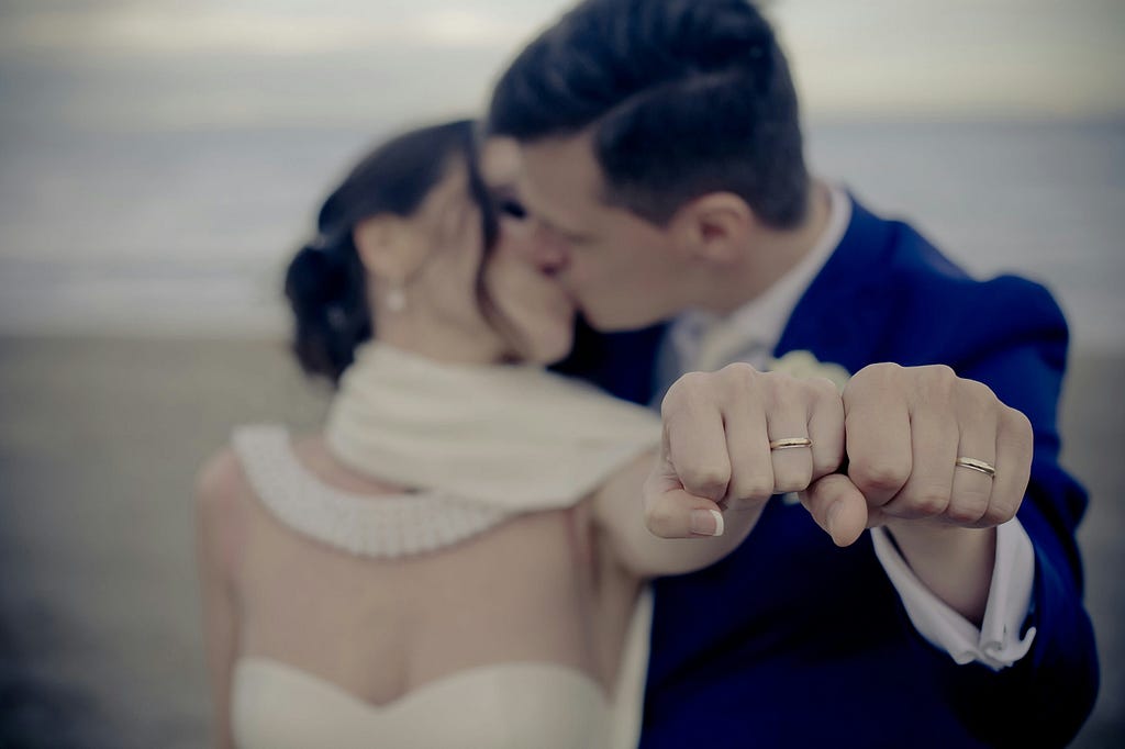 A bride and groom kiss while proudly displaying their custom engagement rings and wedding rings in focus, symbolizing their union.