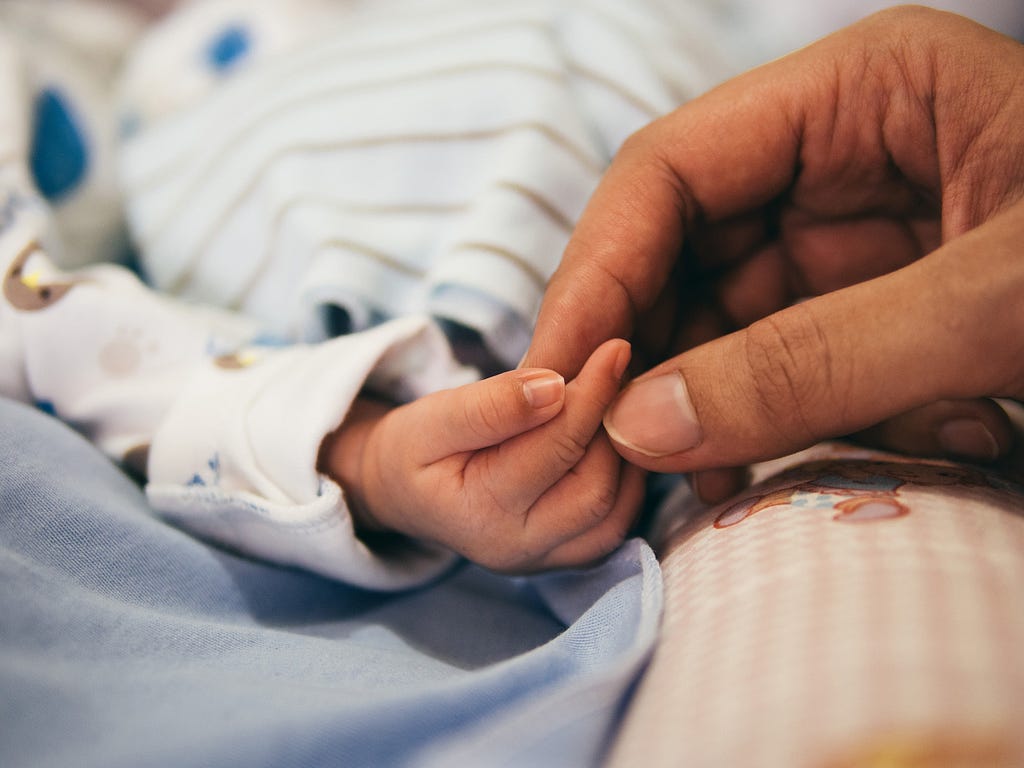 A parent’s index finger and thumb holding their newborn’s index finger. Hospital setting.