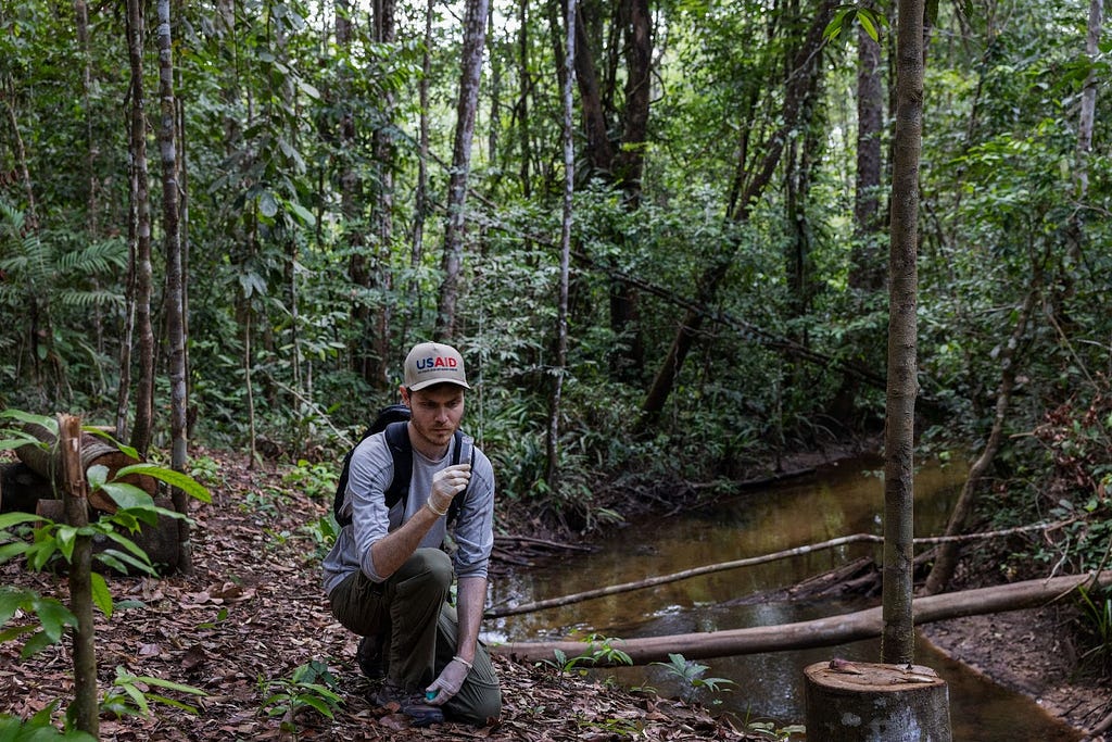 A man wearing gloves and a USAID hat holding a soil sample in the middle of the forest