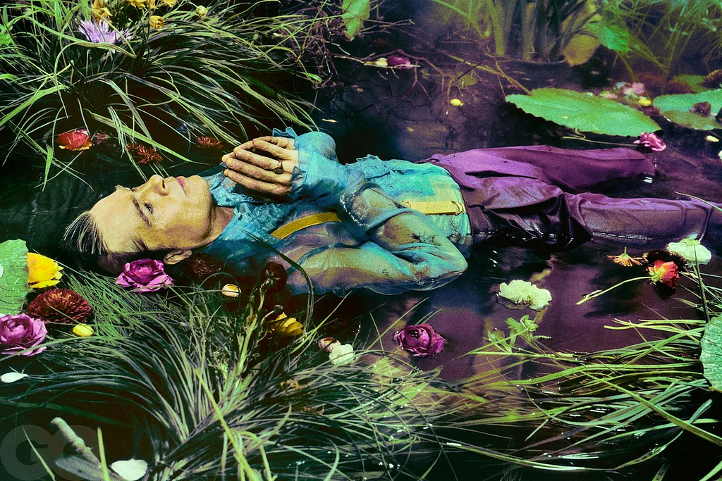 Brad Pitt lays in swampy waters face up with his hands over his chest as if in prayer. He wears a sheer blue short and purple trousers which match the purple flowers and green foliage growing in the water.