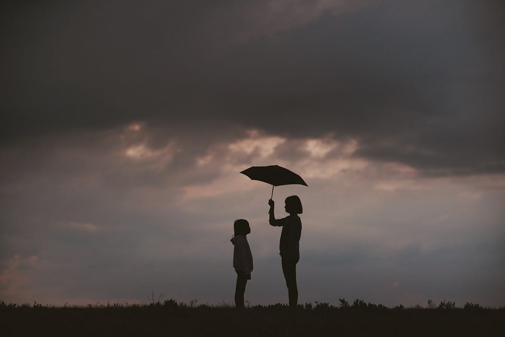 Silhouette of two people (one taller than the other) in the middle of a cloudy landscape. The taller one holds an umbrella.