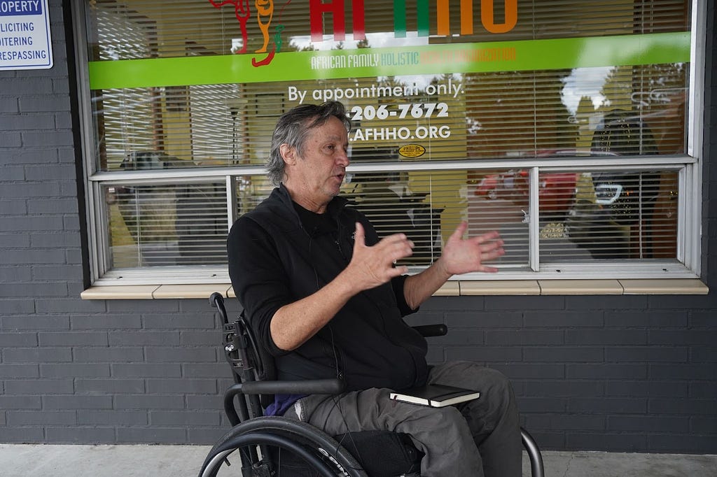 A man seated in a wheel chair gestures in front of an office building window.