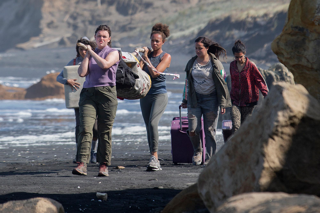 The women settle onto the island, carrying the body of their peer who passed away on the first day on the island.
