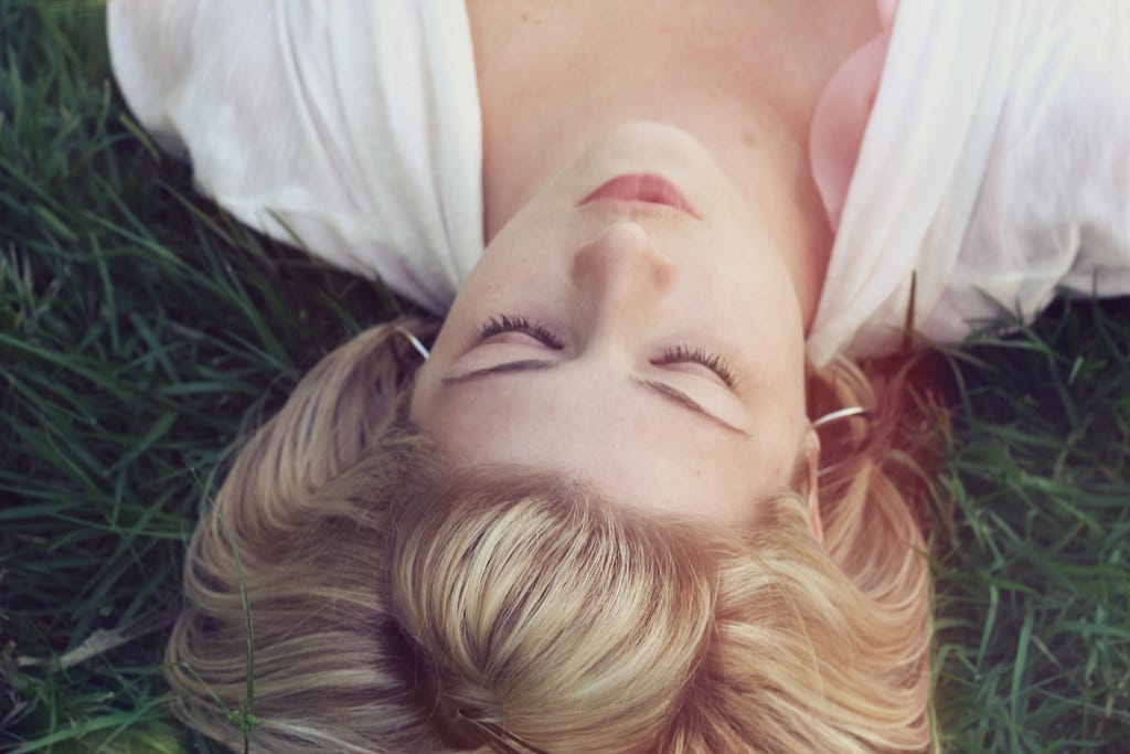 woman with blond hair and white dress laying on her back in the green grass in a state of calmness or quietness as indicated by her closed eyes.