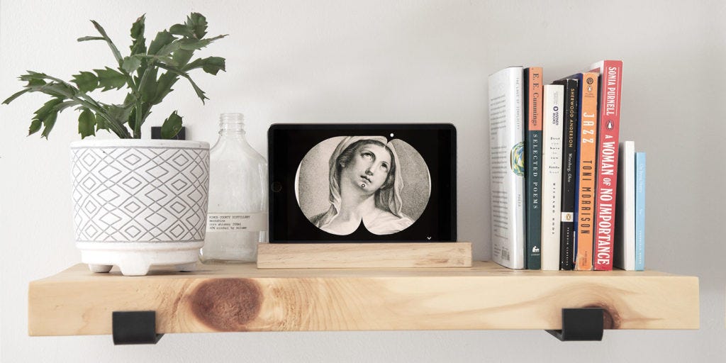 The finished Art Clock displayed on a horizontal tablet sitting on a light wood shelf with books to the right and a Christmas cactus to the left. The Art Clock shows a close cropped etching of a medieval figure with the minute hand pointed to their chin (at 30 past the hour) and the hour hand a 1pm, corresponding to the angle of the figure’s eyes.