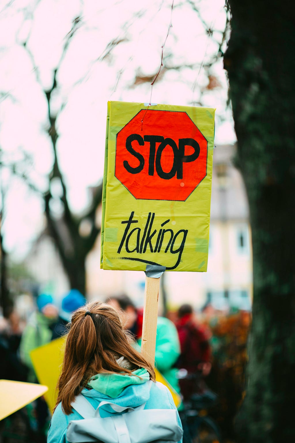 As viewed from behind, a person at a gathering, is holding up a sign. The sign is yellow with an orange-red octagon at the top containing the word “Stop”. Underneath is written “talking”.