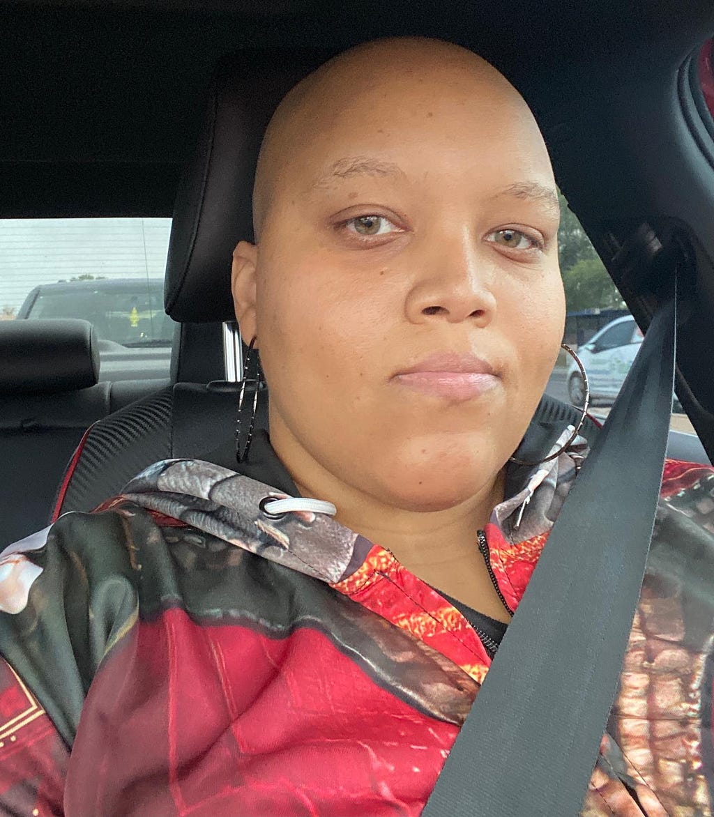 selfie of a light skinned Black woman in a car. She is looking at the camera with a blank expression. Her head is bald, and she’s wearing a hoodie that appears to have a red, brown, and gray pattern in the style of women’s armor.