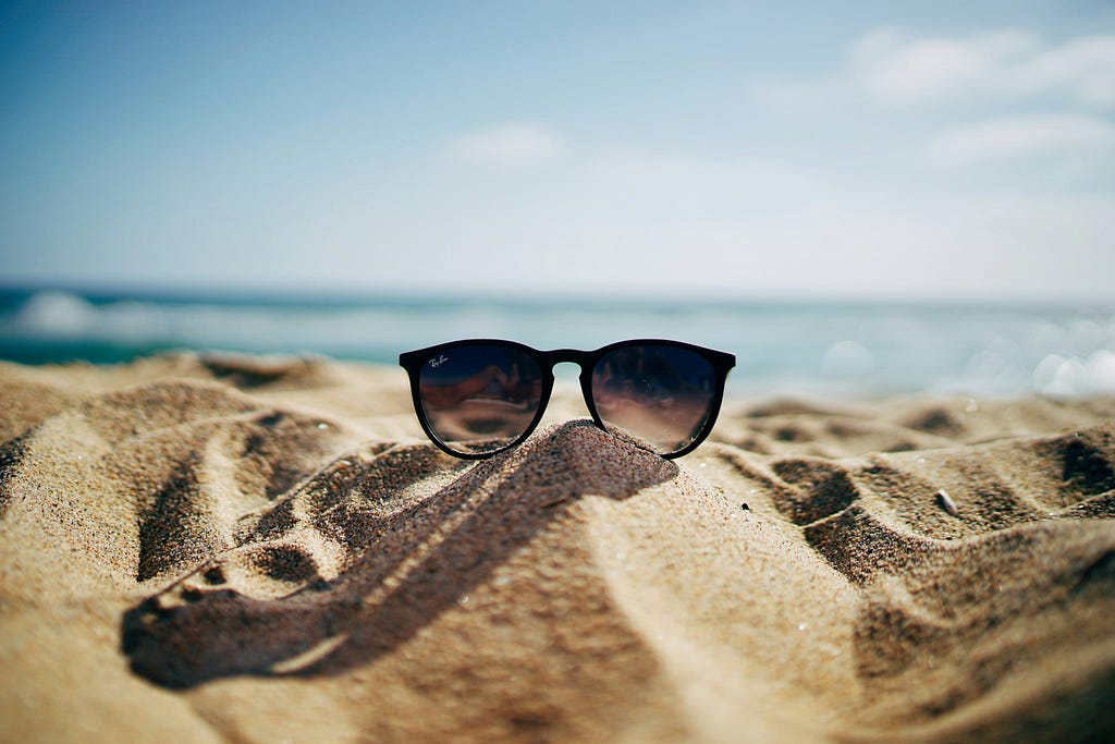 A close up of sunglasses perched on sand with water beyond