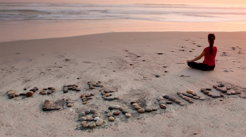 Woman sitting on a beach. The beach has stones arranged on it to say “Work life balance”