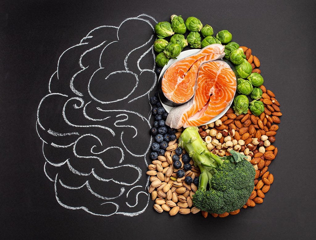 The image shows a drawing of a brain the left just being blank and the right hand side shows different types of food. There are brussle sprouts, salmon, broccoli and a range of nuts such as almonds and pistachio.