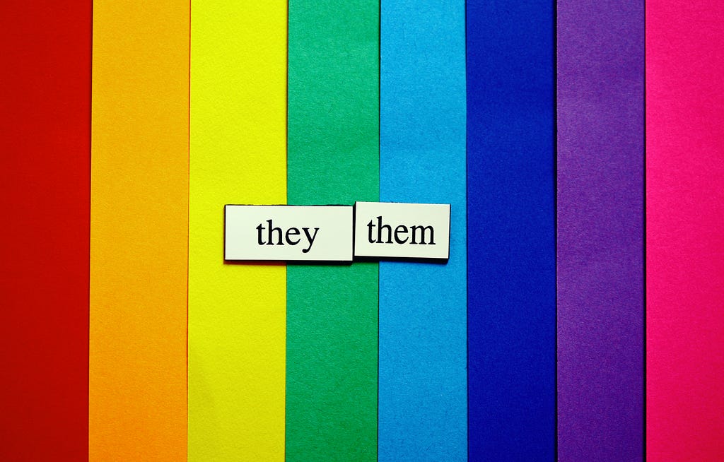 The words “they” and “them” appear over construction paper lined up to show the colors of a rainbow.