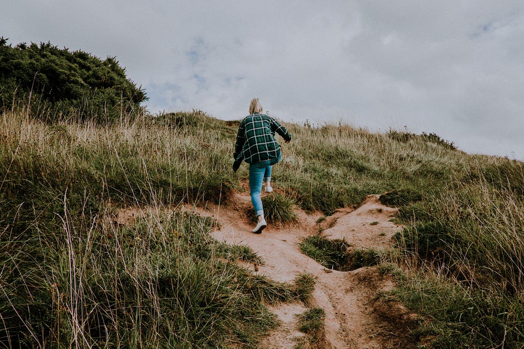 Woman with blonde hair and wearing jeans and a plaid shirt climbs up a sandy, grassy hill. We see her from below — it looks quite steep